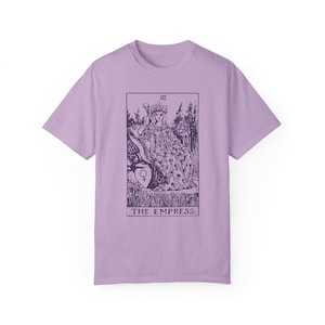 May include: A lavender t-shirt with a graphic of the Empress tarot card. The card features a woman sitting on a throne with a crown on her head. The text "III THE EMPRESS." is printed below the image.
