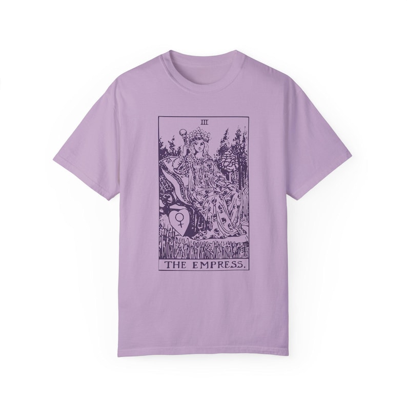 May include: A lavender t-shirt with a graphic of the Empress tarot card. The card features a woman sitting on a throne with a crown on her head. The text "III THE EMPRESS." is printed below the image.