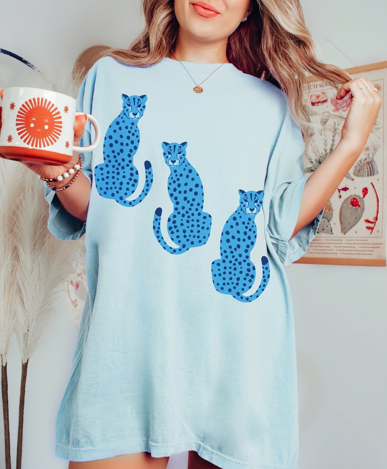 K&ouml;nnte beinhalten: Ein hellblaues Oversize-T-Shirt mit einem grafischen Druck von drei blauen und schwarzen gefleckten Geparden.