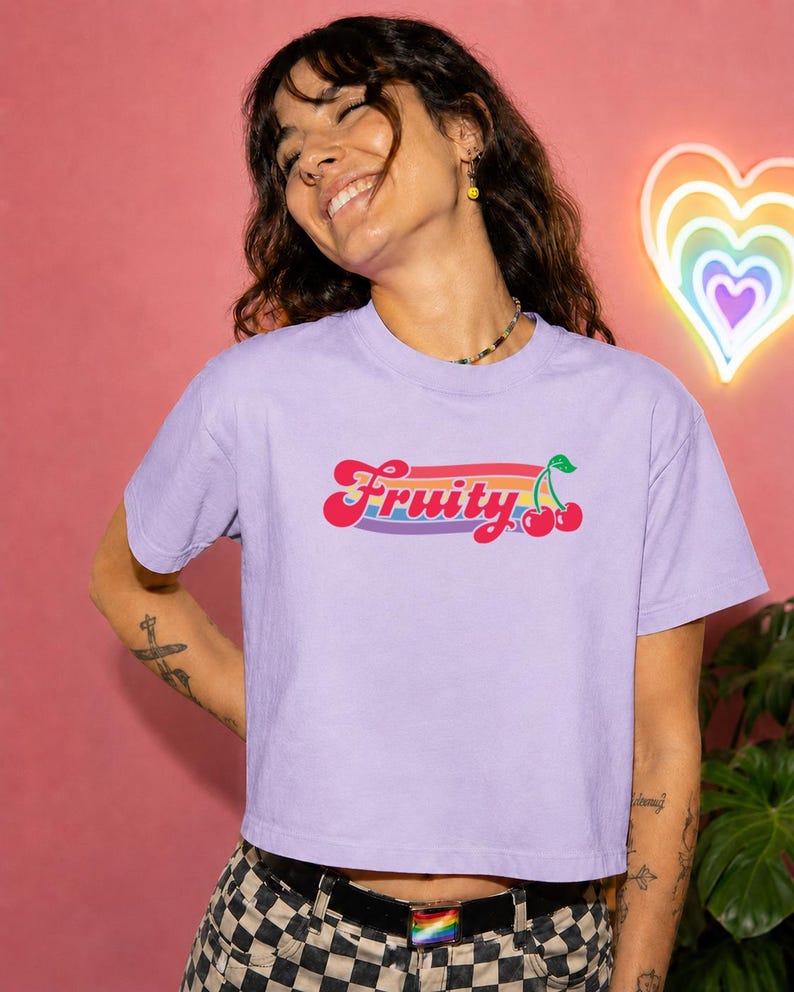 May include: A lilac crop top t-shirt with the word "Fruity" in a retro font, with a rainbow stripe and cherry graphic. The shirt is worn with a black belt and checkered pants. A neon rainbow heart is in the background.