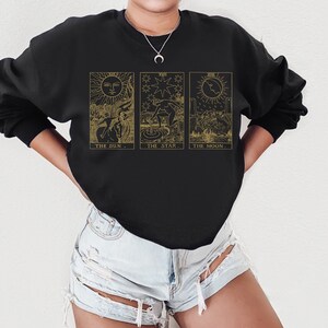 K&ouml;nnte beinhalten: Schwarzes Sweatshirt mit goldenen Tarotkarten-Illustrationen: Die Sonne, Der Stern und Der Mond. Jedes Design hat einen begleitenden Text. Das Sweatshirt ist ein lang&auml;rmeliges Rundhals-Sweatshirt.