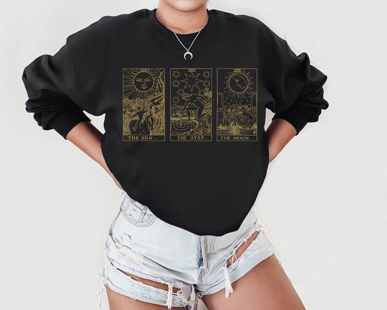 K&ouml;nnte beinhalten: Schwarzes Sweatshirt mit goldenen Tarotkarten-Illustrationen: Die Sonne, Der Stern und Der Mond. Jedes Design hat einen begleitenden Text. Das Sweatshirt ist ein lang&auml;rmeliges Rundhals-Sweatshirt.