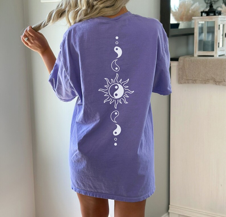Puede incluir: Una camiseta de gran tama&ntilde;o de color morado con un s&iacute;mbolo yin yang blanco dentro de un gr&aacute;fico de sol y s&iacute;mbolos yin yang m&aacute;s peque&ntilde;os arriba y abajo.