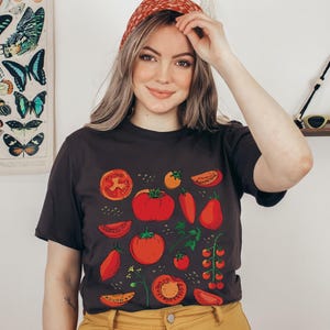 May include: A woman wearing a black t-shirt with a red, green and yellow tomato print. The shirt has a crew neck and short sleeves. The woman is wearing a red headband.