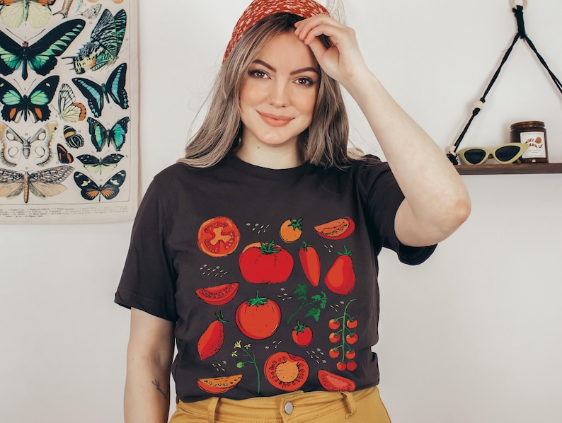 May include: A woman wearing a black t-shirt with a red, green and yellow tomato print. The shirt has a crew neck and short sleeves. The woman is wearing a red headband.