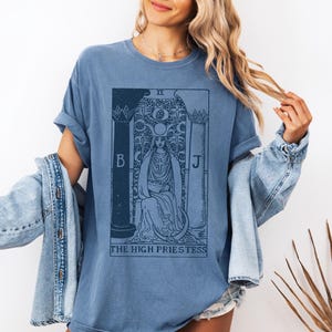 May include: A blue t-shirt with a vintage tarot card design featuring the High Priestess. The card is printed in black and white and includes the Roman numeral II, the letters B and J, and the text "THE HIGH PRIESTESS".