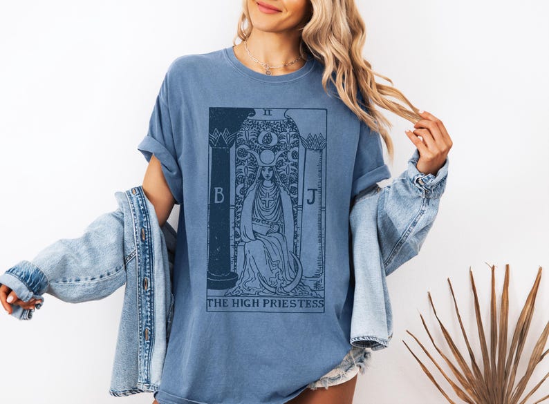 May include: A blue t-shirt with a vintage tarot card design featuring the High Priestess. The card is printed in black and white and includes the Roman numeral II, the letters B and J, and the text "THE HIGH PRIESTESS".