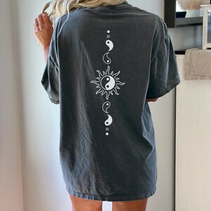 Puede incluir: Una camiseta gris oscura de gran tama&ntilde;o con un s&iacute;mbolo yin yang blanco y un dise&ntilde;o de sol en la espalda.