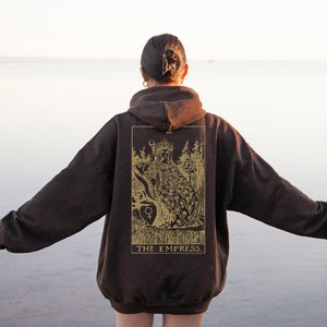 Op de afbeelding: Een donkerbruine hoodie met een goudkleurig tarotkaartontwerp op de rug. Het ontwerp toont een zittende figuur met de tekst "THE EMPRESS". De hoodie is oversized en heeft een capuchon met trekkoord.