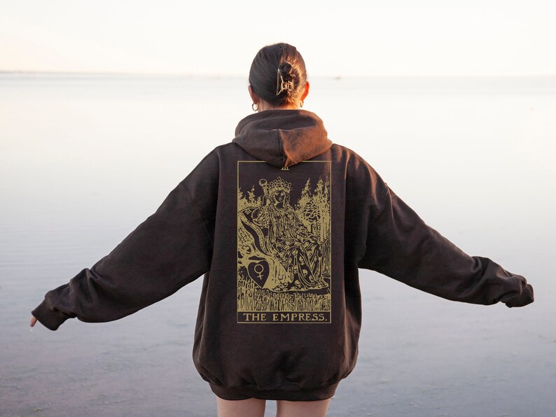 Op de afbeelding: Een donkerbruine hoodie met een goudkleurig tarotkaartontwerp op de rug. Het ontwerp toont een zittende figuur met de tekst "THE EMPRESS". De hoodie is oversized en heeft een capuchon met trekkoord.