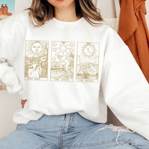 K&ouml;nnte beinhalten: Wei&szlig;es Sweatshirt mit goldenen Tarotkarten-Illustrationen von Sonne, Stern und Mond. Das Sweatshirt hat einen Rundhalsausschnitt und lange &Auml;rmel. Das Design ist mittig auf der Vorderseite, mit den Kartennamen darunter.