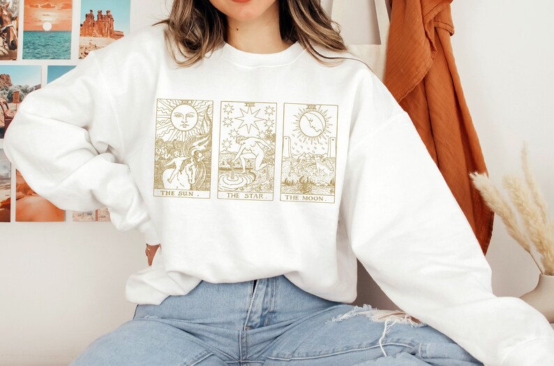 K&ouml;nnte beinhalten: Wei&szlig;es Sweatshirt mit goldenen Tarotkarten-Illustrationen von Sonne, Stern und Mond. Das Sweatshirt hat einen Rundhalsausschnitt und lange &Auml;rmel. Das Design ist mittig auf der Vorderseite, mit den Kartennamen darunter.