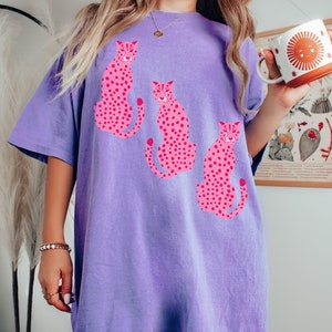 K&ouml;nnte beinhalten: Ein lila T-Shirt mit einem grafischen Druck von drei rosa Leoparden. Die Leoparden sitzen und haben schwarze Flecken.