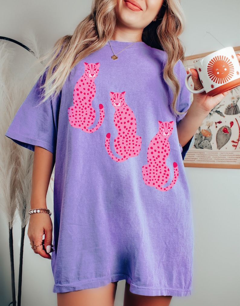 K&ouml;nnte beinhalten: Ein lila T-Shirt mit einem grafischen Druck von drei rosa Leoparden. Die Leoparden sitzen und haben schwarze Flecken.