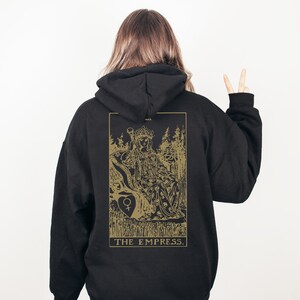 Op de afbeelding: Zwarte hoodie met een gouden illustratie van de tarotkaart De Keizerin op de rug. Het ontwerp toont een zittende figuur, bomen en de tekst "THE EMPRESS."