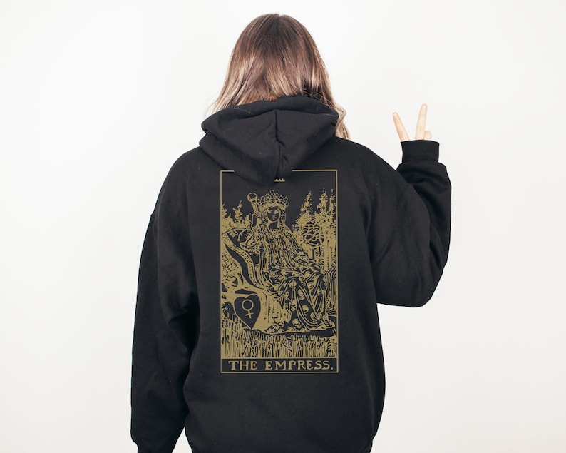 Op de afbeelding: Zwarte hoodie met een gouden illustratie van de tarotkaart De Keizerin op de rug. Het ontwerp toont een zittende figuur, bomen en de tekst "THE EMPRESS."