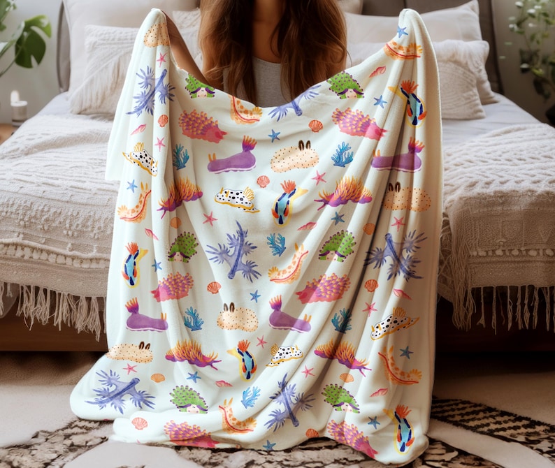 May include: A cream-colored blanket featuring a colorful sea slug pattern. The design includes various sea slugs in shades of purple, orange, green, and blue, along with starfish and coral. The blanket is held up, showcasing the full pattern.