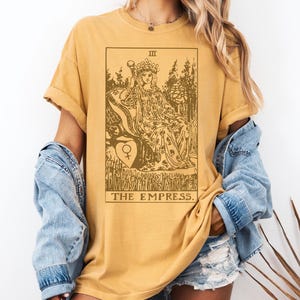 May include: A mustard yellow t-shirt with a brown graphic of the Empress tarot card. The card features a crowned woman sitting on a throne with a symbol of the female gender in a heart.