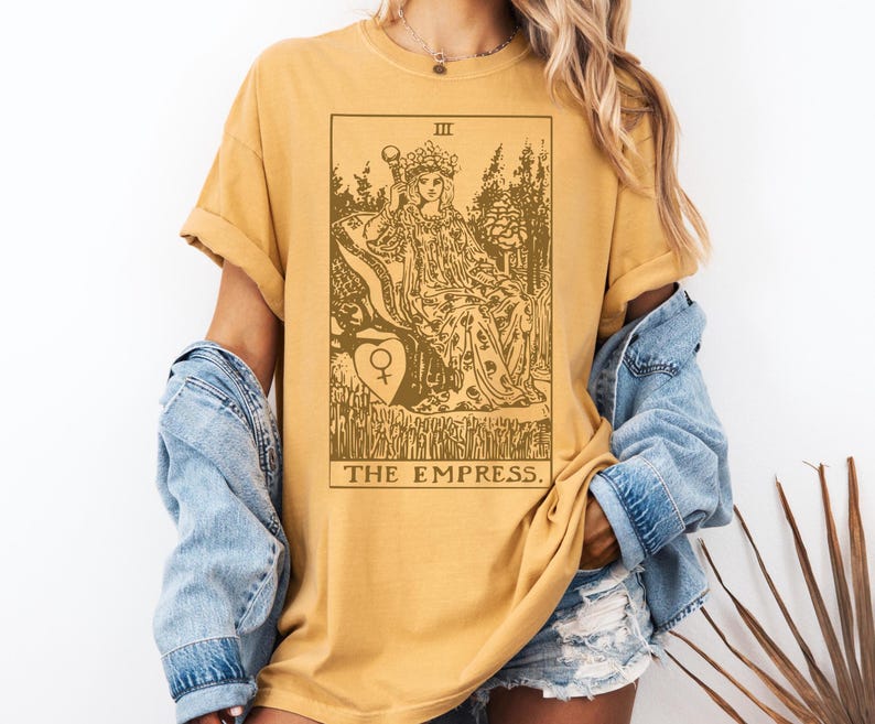 May include: A mustard yellow t-shirt with a brown graphic of the Empress tarot card. The card features a crowned woman sitting on a throne with a symbol of the female gender in a heart.