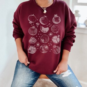 May include: A maroon sweatshirt with a white graphic print of pomegranates. The pomegranates are drawn in a vintage style and are scattered across the front of the sweatshirt.