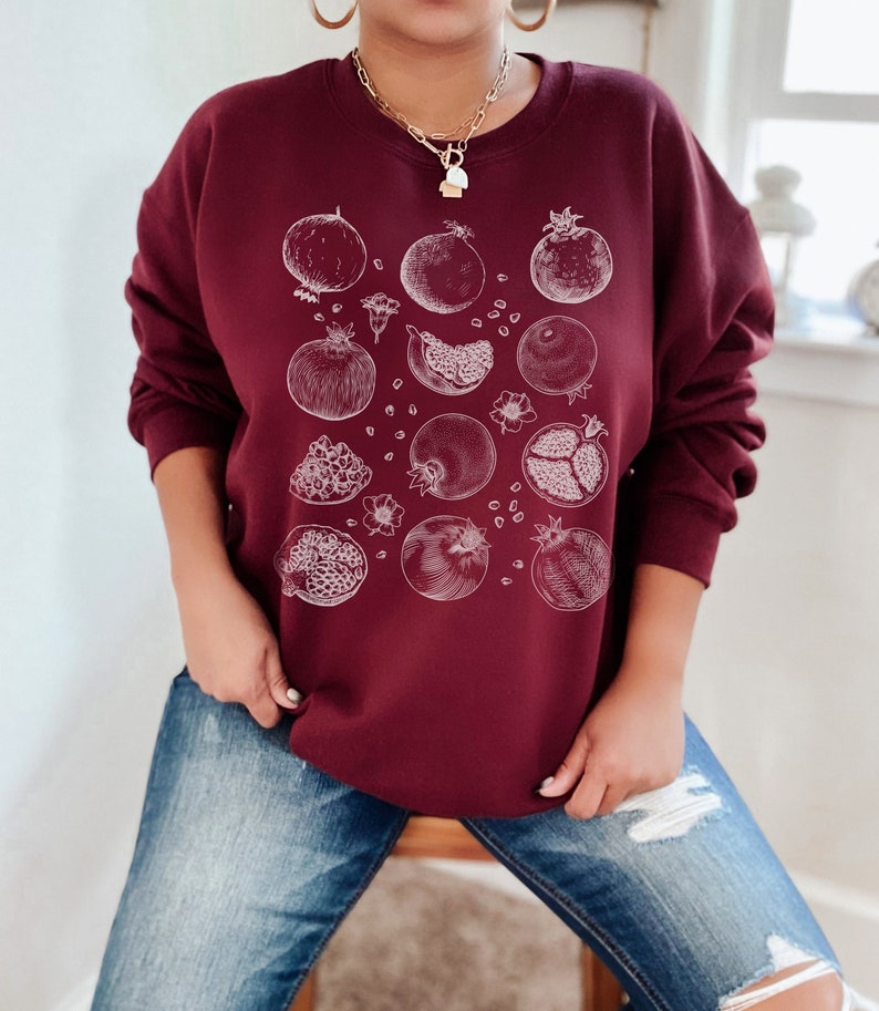 May include: A maroon sweatshirt with a white graphic print of pomegranates. The pomegranates are drawn in a vintage style and are scattered across the front of the sweatshirt.