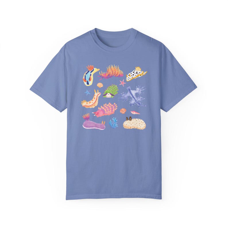 Pu&ograve; includere: Una maglietta blu chiaro con un'illustrazione colorata di varie lumache di mare, note anche come nudibranchi, su uno sfondo bianco. Le lumache di mare sono raffigurate in vari colori, tra cui rosa, viola, blu, giallo e arancione.