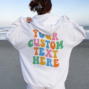May include: White hoodie with a colorful "YOUR CUSTOM TEXT HERE" graphic on the back. The hoodie is worn by a person standing on a sandy beach.