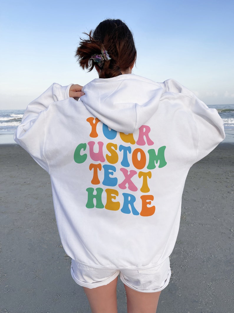 May include: White hoodie with a colorful "YOUR CUSTOM TEXT HERE" graphic on the back. The hoodie is worn by a person standing on a sandy beach.