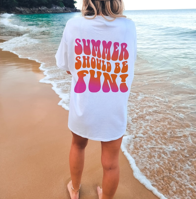 以下が含まれることがあります： レトロな波型のフォントで「SUMMER SHOULD BE FUN！」と書かれた白いTシャツ。テキストはピンクからオレンジに変化します。Tシャツは、海を背景にしたビーチで着用されています。
