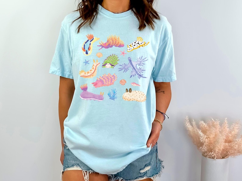 Pu&ograve; includere: Una maglietta blu chiaro con una stampa grafica colorata di varie lumache di mare. La grafica presenta una variet&agrave; di colori e forme, tra cui rosa, viola, blu, verde e giallo.