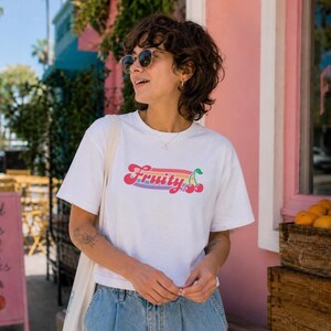 May include: White t-shirt with the word "Fruity" in a colorful, retro font, featuring cherries. The shirt is worn by a person wearing sunglasses and blue jeans. A sign nearby says "good vibes cold drinks".