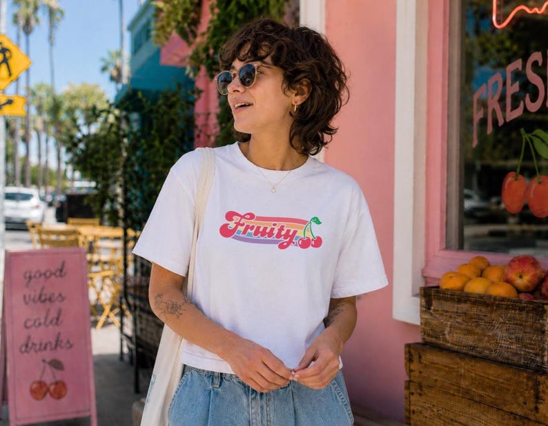 May include: White t-shirt with the word "Fruity" in a colorful, retro font, featuring cherries. The shirt is worn by a person wearing sunglasses and blue jeans. A sign nearby says "good vibes cold drinks".