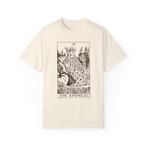 May include: A beige t-shirt with a brown graphic of the Empress tarot card. The card features a woman sitting on a throne with a crown and a scepter. The text "III THE EMPRESS." is printed below the card.