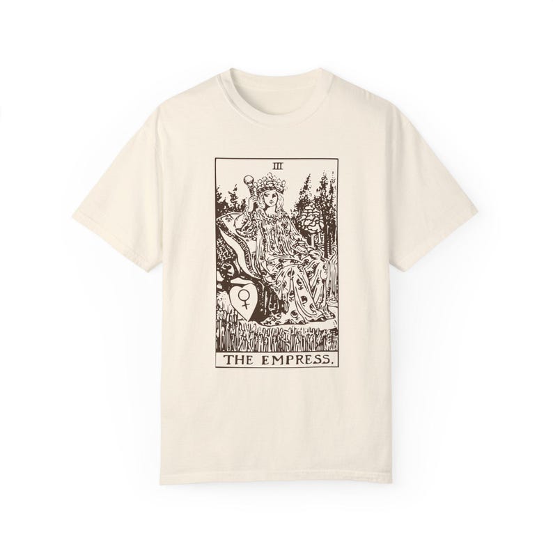 May include: A beige t-shirt with a brown graphic of the Empress tarot card. The card features a woman sitting on a throne with a crown and a scepter. The text "III THE EMPRESS." is printed below the card.