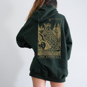 Op de afbeelding: Donkergroene hoodie met een goudkleurige afbeelding van de tarotkaart De Keizerin op de rug. Het ontwerp bevat een illustratie van een zittende figuur, bomen en de titel van de kaart. De hoodie heeft een capuchon met trekkoord en een kangoeroezak.