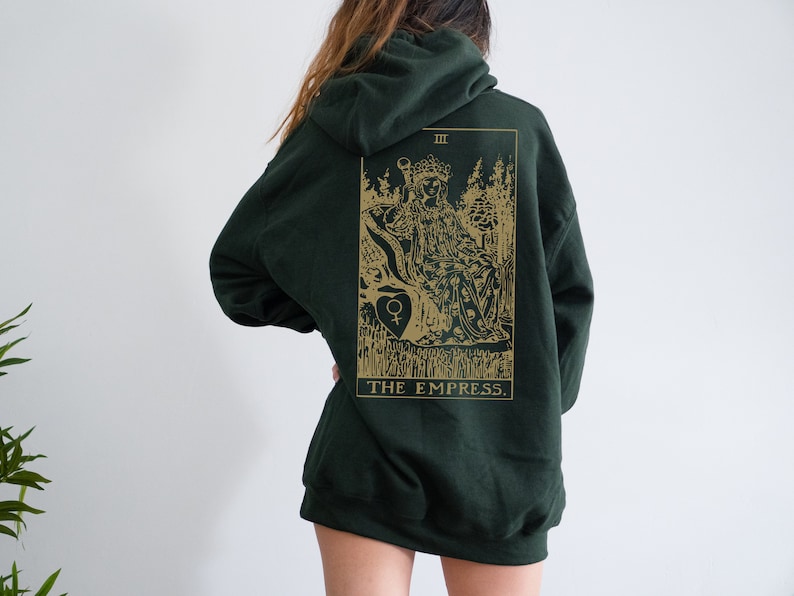 Op de afbeelding: Donkergroene hoodie met een goudkleurige afbeelding van de tarotkaart De Keizerin op de rug. Het ontwerp bevat een illustratie van een zittende figuur, bomen en de titel van de kaart. De hoodie heeft een capuchon met trekkoord en een kangoeroezak.