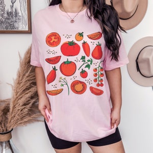 May include: A pink t-shirt featuring a graphic print of red tomatoes, green leaves, and tomato seeds. The design includes various shapes and sizes of tomatoes, such as whole tomatoes, slices, and halved tomatoes.