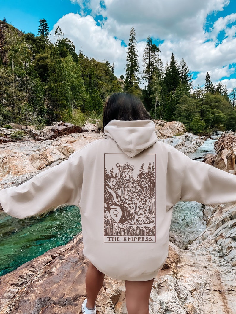 Op de afbeelding: Beige hoodie met een capuchon en een grote afbeelding op de rug. De afbeelding is een bruine illustratie van een tarotkaart, "The Empress". De hoodie wordt buiten gedragen in de buurt van een rivier.