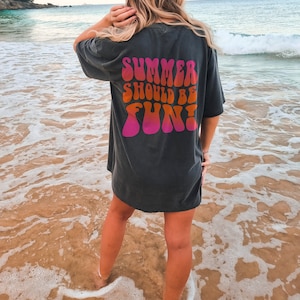 以下が含まれることがあります： レトロな波型のフォントで「SUMMER SHOULD BE FUN！」と書かれた濃いグレーのTシャツ。テキストはピンク、オレンジ、赤の色合いです。Tシャツはビーチで着用されています。