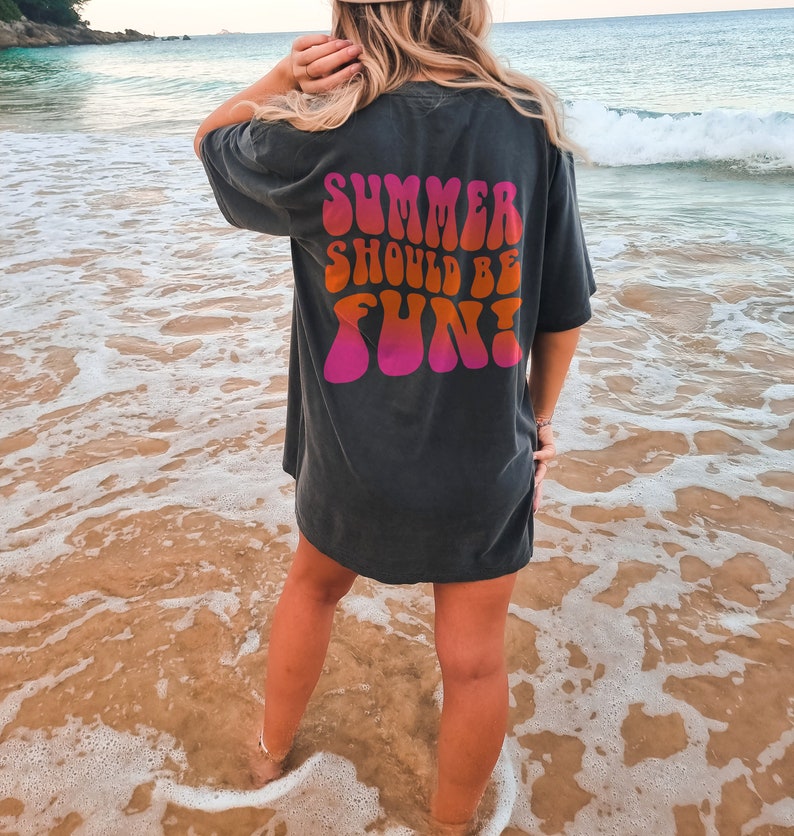 以下が含まれることがあります： レトロな波型のフォントで「SUMMER SHOULD BE FUN！」と書かれた濃いグレーのTシャツ。テキストはピンク、オレンジ、赤の色合いです。Tシャツはビーチで着用されています。