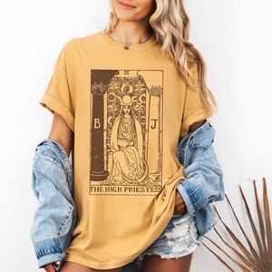 May include: A mustard yellow t-shirt with a brown graphic of the High Priestess tarot card. The card features a woman sitting on a throne with a crescent moon above her head. The text "The High Priestess" is printed below the card.