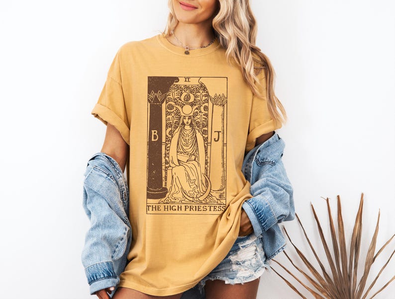 May include: A mustard yellow t-shirt with a brown graphic of the High Priestess tarot card. The card features a woman sitting on a throne with a crescent moon above her head. The text "The High Priestess" is printed below the card.