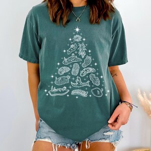 May include: Teal-colored t-shirt featuring a white graphic of a Christmas tree made of sea slugs and other marine life. The design includes a star at the top and small stars throughout the tree shape. The shirt has short sleeves and a relaxed fit.