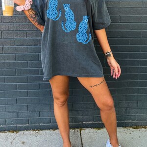K&ouml;nnte beinhalten: Ein &uuml;berdimensioniertes, dunkelgraues T-Shirt mit einem grafischen Druck von drei blauen und wei&szlig;en Geparden-Silhouetten. Das Shirt hat eine lockere Passform und kurze &Auml;rmel.