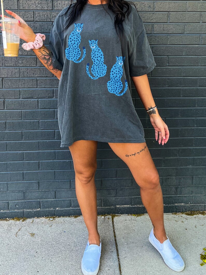 K&ouml;nnte beinhalten: Ein &uuml;berdimensioniertes, dunkelgraues T-Shirt mit einem grafischen Druck von drei blauen und wei&szlig;en Geparden-Silhouetten. Das Shirt hat eine lockere Passform und kurze &Auml;rmel.