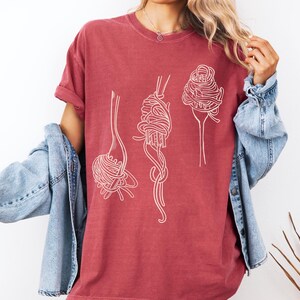 K&ouml;nnte beinhalten: Ein maronfarbenes T-Shirt mit einer wei&szlig;en Umrandung von Spaghetti auf Gabeln. Das Shirt hat eine lockere Passform und einen Rundhalsausschnitt.