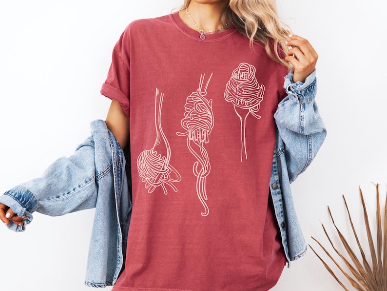 K&ouml;nnte beinhalten: Ein maronfarbenes T-Shirt mit einer wei&szlig;en Umrandung von Spaghetti auf Gabeln. Das Shirt hat eine lockere Passform und einen Rundhalsausschnitt.