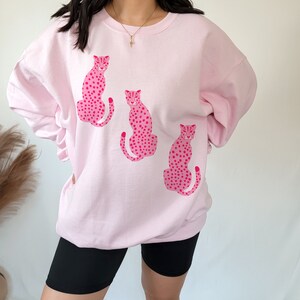 May include: Pink sweatshirt with a graphic print of three pink leopards with black spots.