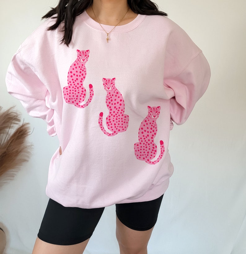 May include: Pink sweatshirt with a graphic print of three pink leopards with black spots.