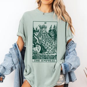 May include: A light green t-shirt with a graphic of the Empress tarot card. The card features a woman sitting on a throne with a crown on her head. The text "THE EMPRESS" is printed below the card.
